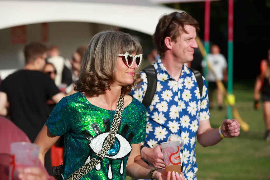 Two people among an outdoor crowd, one wearing heart-shaped sunglasses and a green sequinned top.