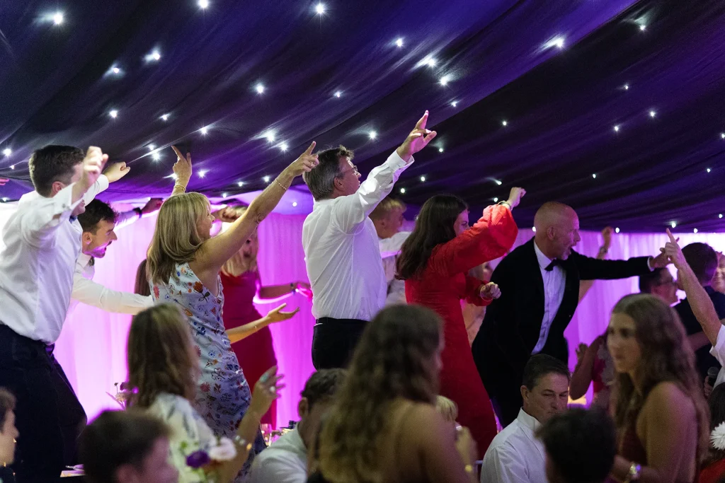 Standing guests stretch their arms upwards beneath a canopy of small white lights