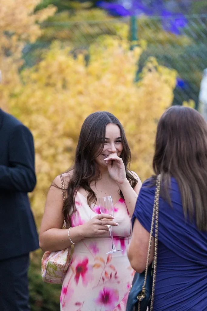 Two guests holding drinks face one another in the garden, with one smiling behind her hand