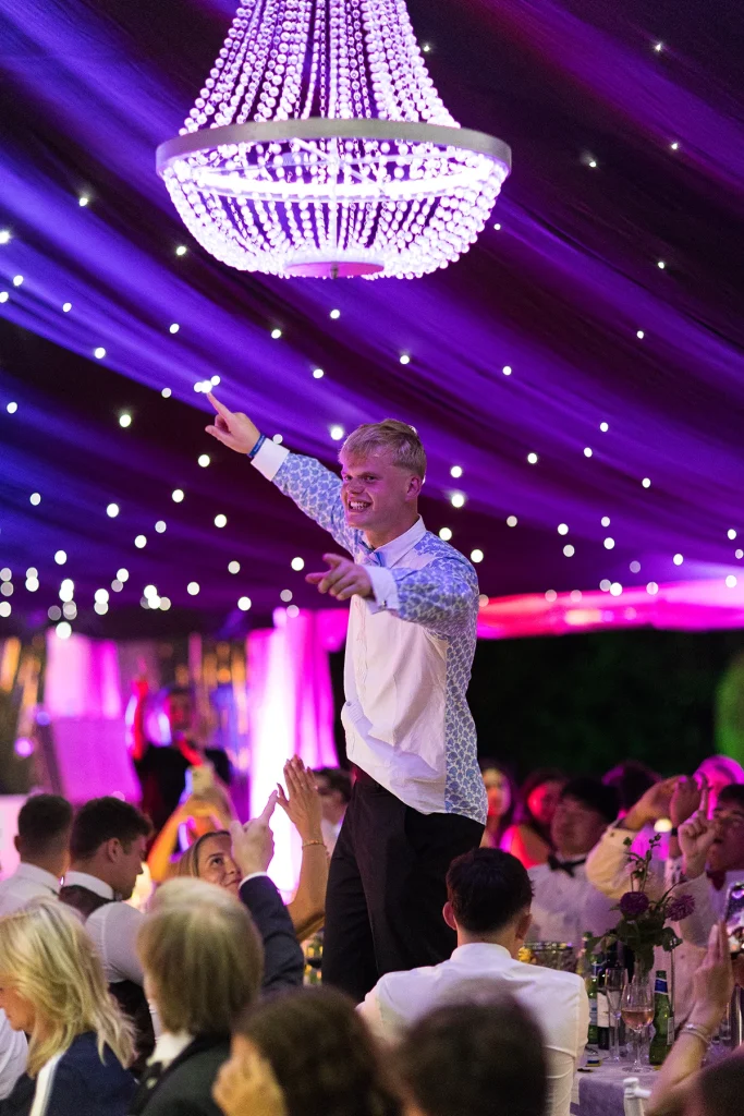 A smiling guest stands above the seated gathering with arms outstretched beneath a chandelier