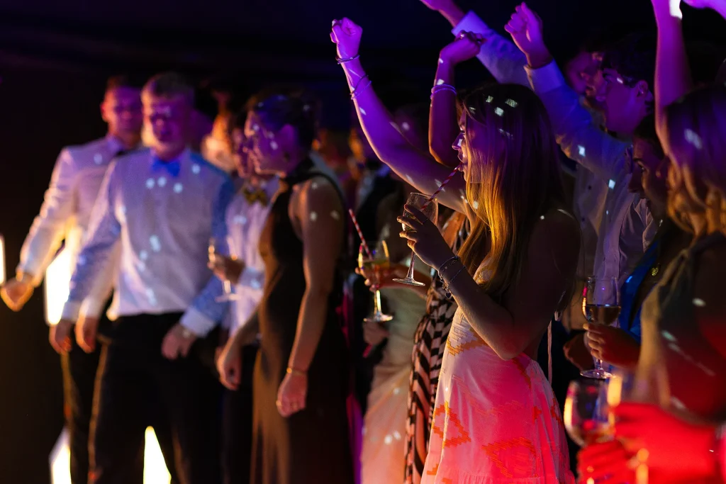 Guests holding drinks dance with their arms raised under coloured evening lighting