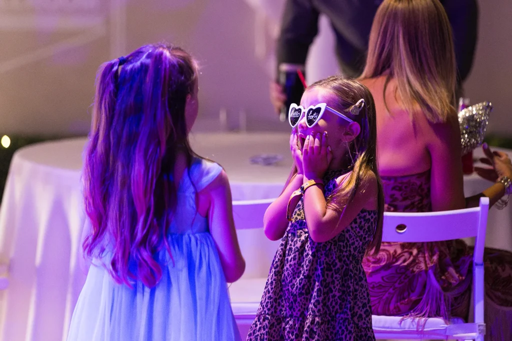 Two girls facing one another, one wearing heart-shaped party glasses with her hands against her cheeks