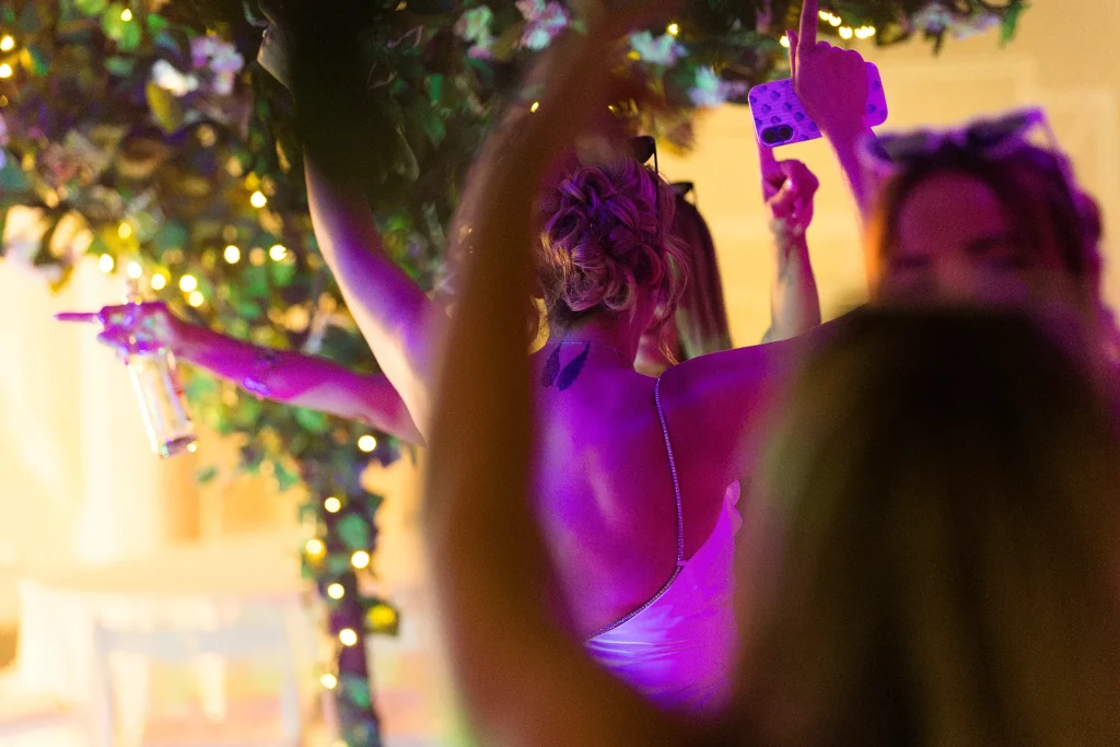 Guests raising their arms beneath foliage and coloured lights, with a phone held aloft