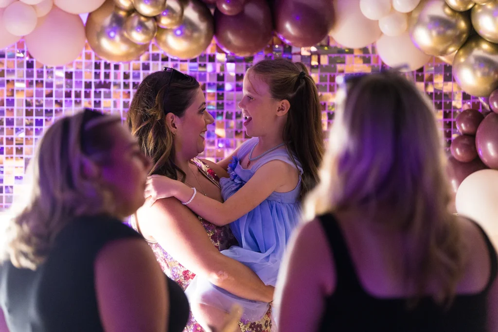 A woman holding a smiling girl in front of balloons and a reflective backdrop, with guests in the foreground