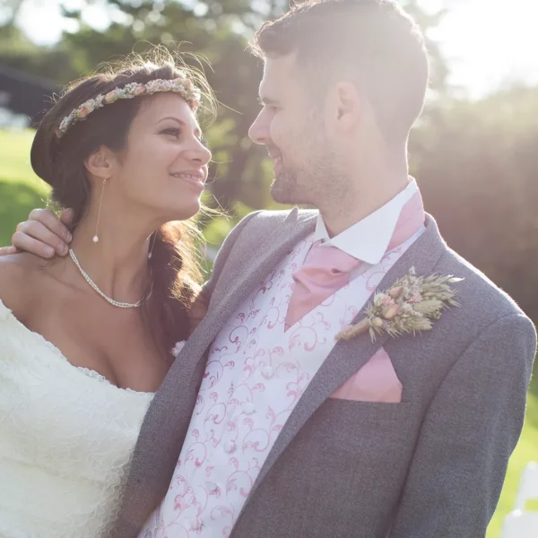 Bride and groom smiling at each other outdoors with bright sunshine behind them